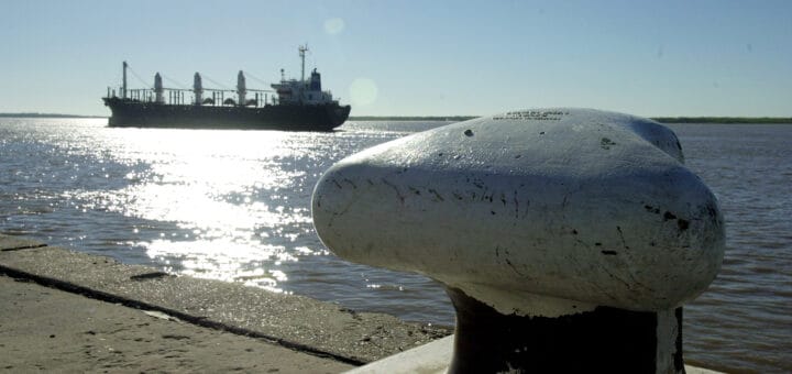 alt="en la imagen puede verse un barco navegando por un río, en primer plano se observa parte de un puerto"