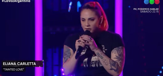 alt="una mujer, con tatuajes en los brazos, remera negra y un micrófono sostenido entre sus manos"