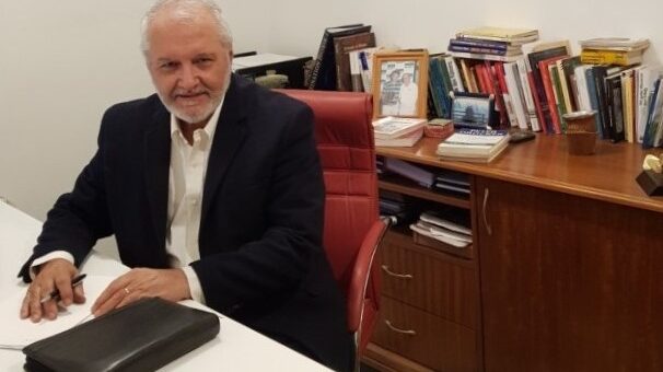 alt="un hombre de traje oscuro y camisa blanca, de cabello canoso, sentado frente a un escritorio, detrás un mueble con libros en su parte superior"
