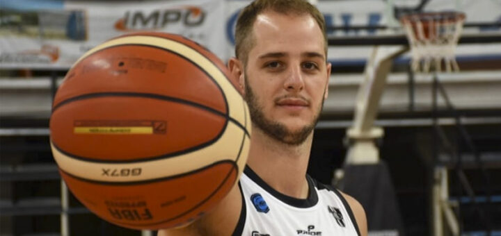 alt="en la fotografía, un jugador de básquet sostiene una pelota en su mano derecha, detrás un aro y parte de un estadio"