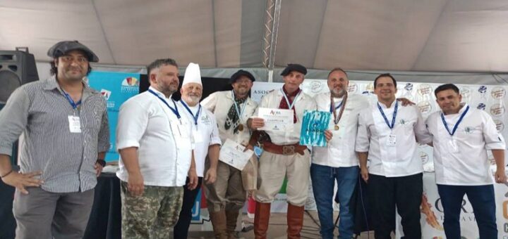 alt="en el centro de la foto, con botas, bombacha, boina, camisa y pañuelo al cuello, los campeones exhiben sus certificados, rodeados de chefs y otros asadores"