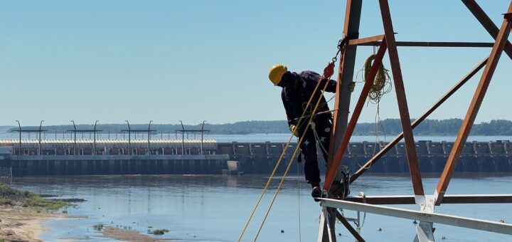 alt="Operarios instalando equipos sobre una torre metálica para la prueba de sirenas de Salto Grande con el río y la represa de fondo"