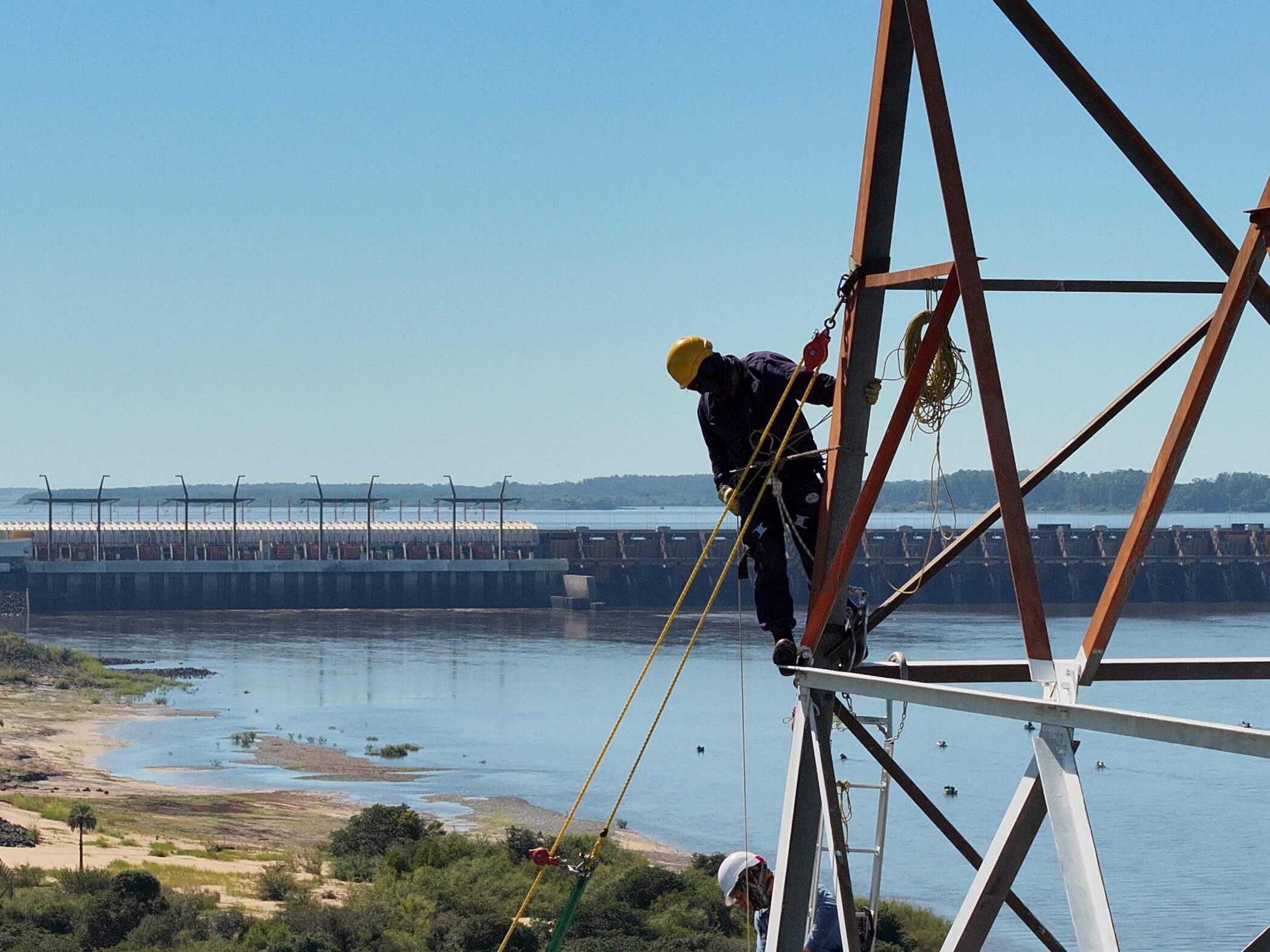 alt="Operarios instalando equipos sobre una torre metálica para la prueba de sirenas de Salto Grande con el río y la represa de fondo"