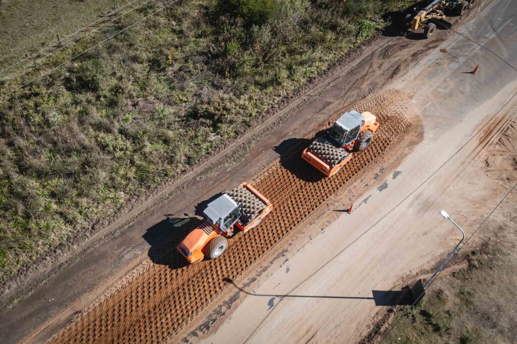 alt="Vista aérea cenital y recta de dos aplanadoras viales trabajando en fila sobre un tramo largo de tierra marrón al lado de un sector pavimentado. Se distinguen las sombras de las maquinarias proyectadas sobre el asfalto por la luz del sol"