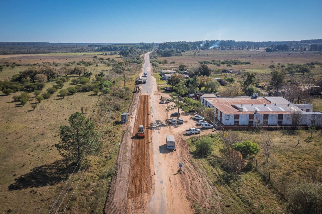 alt="Fotografía aérea de un tramo recto de ruta provincial 4 en medio de campos verdes de Entre Ríos. Un carril de la ruta se encuentra intervenido con tierra marrón removida y una máquina compactadora en actividad. Al costado derecho se observa un complejo de edificaciones rurales con vehículos estacionados"