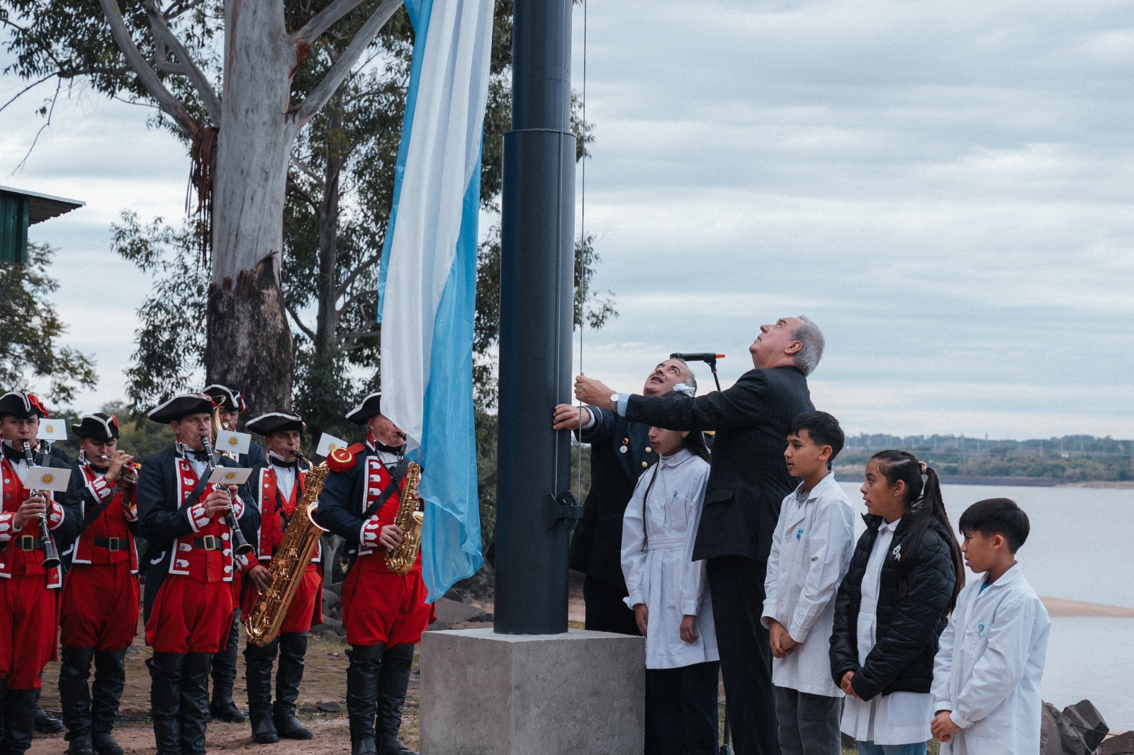 alt="Izamiento de la bandera nacional junto al lago en el acto del 25 de Mayo en Salto Grande con presencia de escolares y autoridades"