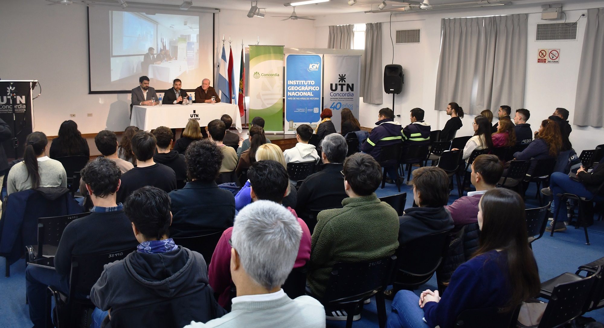 alt="Vista de sala llena en la apertura de las capacitaciones SIG-IDE en Concordia, con expositores y banderas institucionales"