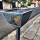 alt="Canasto metálico negro para la disposición y recolección de residuos en Concordia, ubicado sobre una vereda en un día despejado"