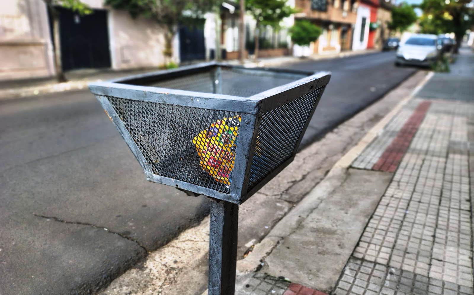 alt="Canasto metálico negro para la disposición y recolección de residuos en Concordia, ubicado sobre una vereda en un día despejado"